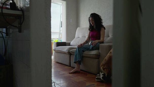 Задумчивая женщина, сидящая на диване с собакой, гуляющей по квартире. Непринужденный быт - Фото, изображение
