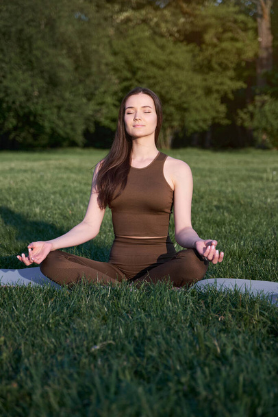 A young woman sits in a lotus position and meditates in the park on a warm summer day. The concept of a healthy lifestyle. - Zdjęcie, obraz