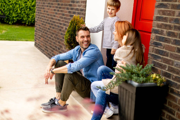Family with a mother, father, son and daughter sitting outside on steps of a front porch of a brick house - Foto, afbeelding
