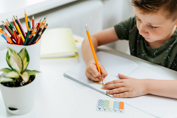 Школьник учится дома с книгой, рисует блокнот и делает домашнее задание. Думаю, кавказский ребенок сидит за столом с блокнотом. Возвращайся в школу. Дистанционное обучение онлайн. - Фото, изображение