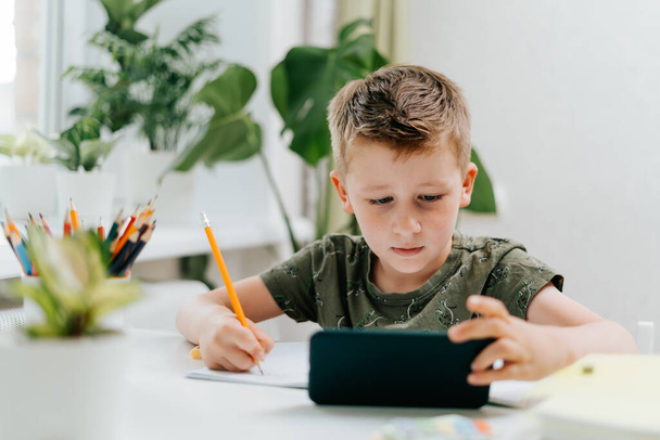 School kid boy studying at home with mobile smart phone and doing homework. Back to school. Thinking caucasian child with cellphone, pencil and training books. Distance learning online education. - Photo, Image