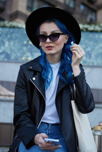 Stylish young woman in black leather jacket and hipster hat listening to music outdoor. Portait of a beautiful diverse female person with dyed blue hair using wireless headphones and a smart phone - Foto, Imagem