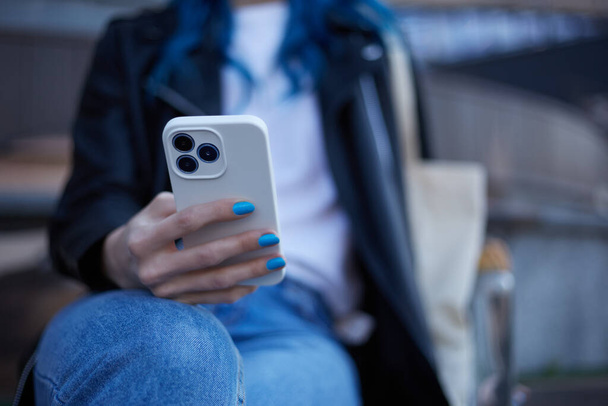 Young woman browsing a mobile app on a modern smart phone with triple camera. Unrecognizable female person sitting on a bench and using a cell phone - 写真・画像