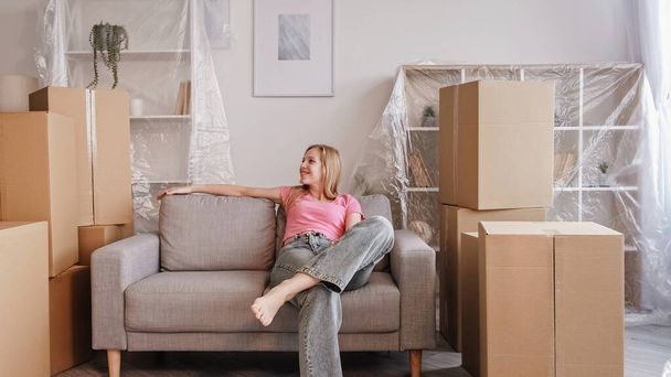 Moving house. Home relocation. Happy smiling young woman sitting on sofa enjoying new living place surrounded by boxes and covered furniture. - Photo, Image