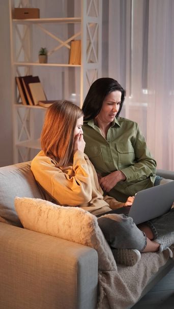Internet disgust. Family browsing. Woman teenage daughter watching scary video on laptop online evening together network sitting sofa light shadow room interior. - Foto, imagen