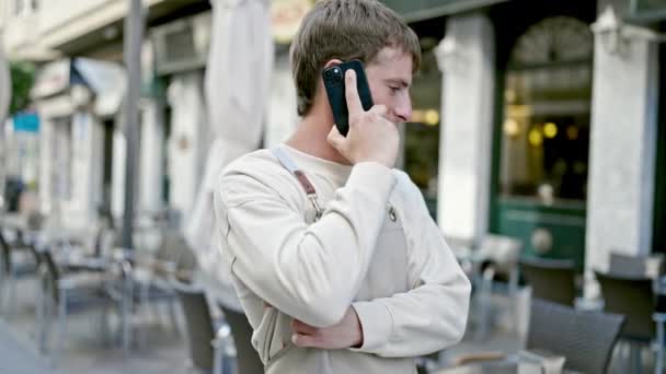 Fiatal kaukázusi férfi pincér mosolygós magabiztos beszél okostelefon kávézóban terasz - Felvétel, videó