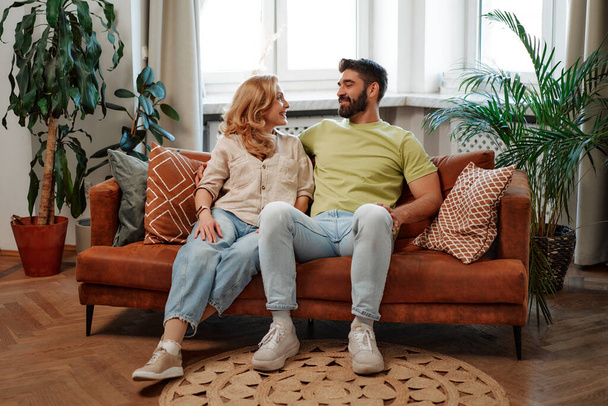 Beautiful adult couple hugging, sitting on the sofa by the window in the living room at home, chatting, spending time together romantically. - Photo, Image