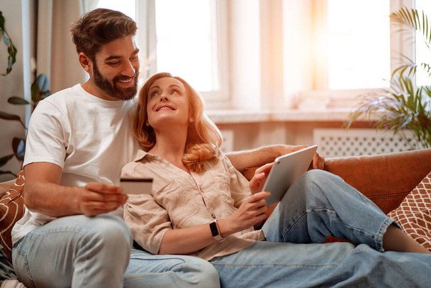 Gyönyörű felnőtt pár fekszik a kanapén tabletta és hitelkártya, csinál online vásárlás a nappaliban otthon, csevegés, időt töltenek együtt romantikusan. - Fotó, kép