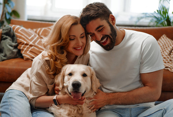 Young couple playing and hugging their pet labrador dog while sitting on the sofa by the window in the living room at home, having fun together. - Photo, Image