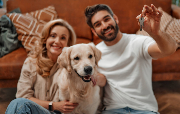 Mudar de casa para comprar um apartamento novo. Adulto casal apaixonado por seu cão labrador, segurando as chaves da casa, sentado no chão perto do sofá na sala de estar no novo apartamento. - Foto, Imagem