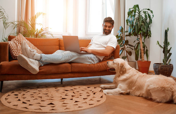 Jóképű szakállas férfi ül a kanapén ablak mellett laptop mellett labrador kutya feküdt a padlón, miközben szabadúszó online vásárlás vagy csevegés a nappaliban otthon. - Fotó, kép