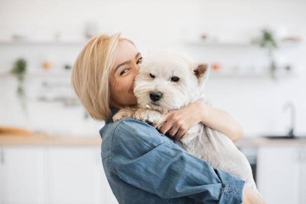 Portrait of smiling blonde lady with West Highland White Terrier in arms posing on background of spacious kitchen. Happy caucasian pet owner rewarding good behavior during day with gentle hug. - Foto, Bild