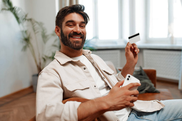 Yetişkin sakallı yakışıklı bir adam oturma odasında arka plandaki koltukta oturuyor, elinde bir kredi kartı ve akıllı bir telefon var. İnternetten alışveriş. - Fotoğraf, Görsel