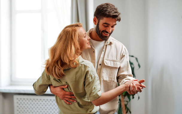 An adult couple in love dancing merrily in the living room against the background of the sofa and the window at home, having fun and romantic time together. - Foto, imagen