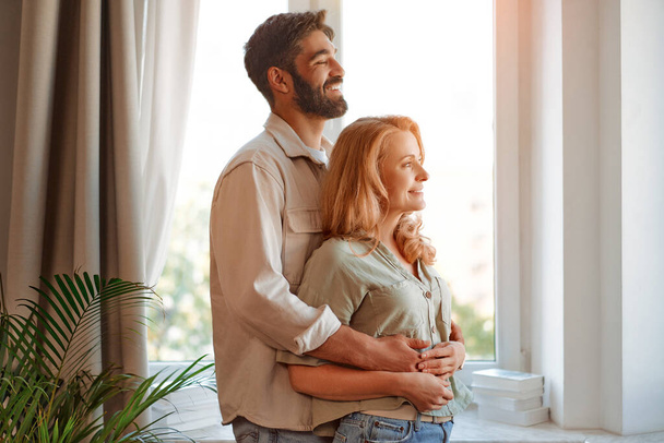 Όμορφο ζευγάρι ενηλίκων αγκαλιάζει ενώ στέκεται από το παράθυρο στο σαλόνι στο σπίτι, περνώντας χρόνο μαζί ρομαντικά. - Φωτογραφία, εικόνα
