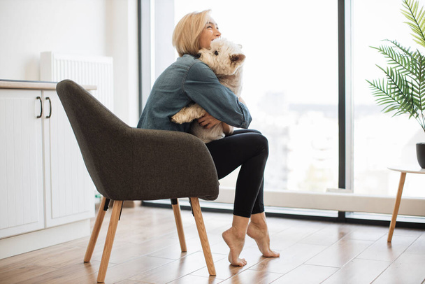 Beautiful slim woman looking at cute dog nose to nose while staying in armchair with feet standing on toes. Playful short-haired female keeper laughing together with canine buddy on weekend at home. - Fotografie, Obrázek
