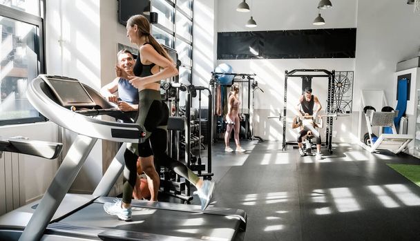 Full body of cheerful young female athlete in sportswear exercising on treadmill while talking to male instructor during fitness workout in modern gym - Foto, imagen