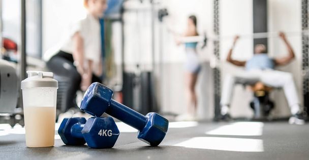 Ground level of blue dumbbells with bottle of protein drink placed on floor near unrecognizable male and female athletes in modern gym on sunny day - Foto, afbeelding