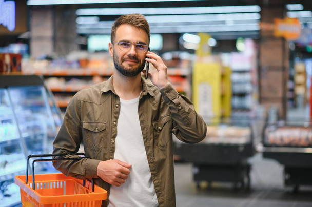 Happy man buying healthy food and drink in a modern supermarket and talking on phone - Foto, Imagen