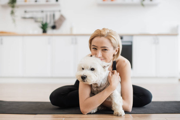 Affectionate adult female in yoga activewear embracing white terrier while sitting cross-legged on rubber mat indoors. Healthy lady awarding sweet pet with hug for good behaviour during home workout. - Photo, image