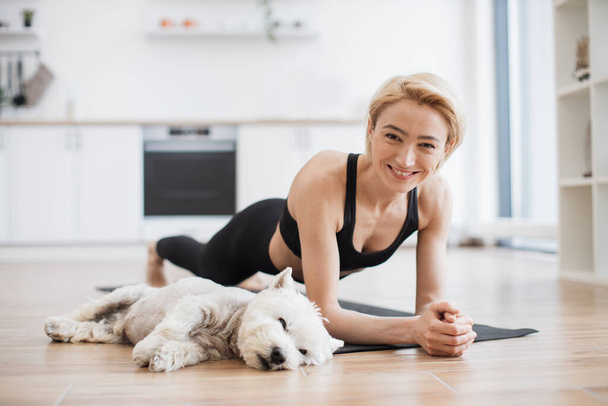 Peaceful blonde lady in black activewear doing yoga practice while tired Westie resting beside on floor of kitchen. Athletic yogini holding Forearm Plank Pose during yoga session with pet at home. - Foto, Imagem