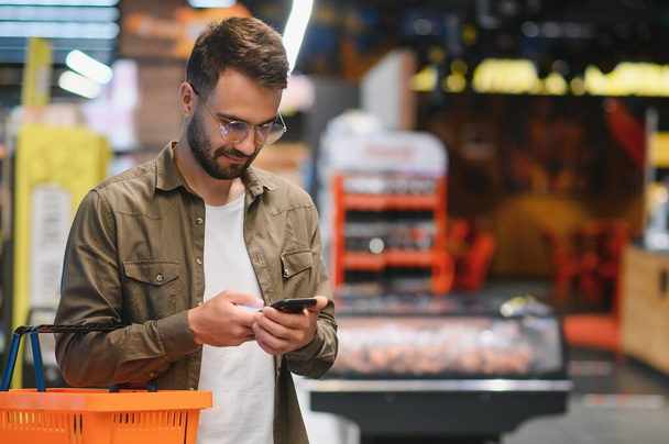 At the Supermarket: Handsome Man Uses Smartphone and Looks at Nutritional Value of the Canned Goods. He's Standing with Shopping Cart in Canned Goods Section - Фото, изображение