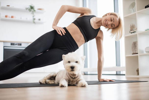 Selective view of attractive slim adult in activewear working out Vasisthasana exercise while small pet relaxing on floor. Good-natured Westie becoming well-suited buddy for yoga workout indoors. - Foto, Bild