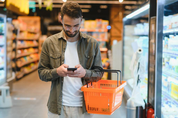 Quick text during shopping. Handsome young man holding mobile phone and smiling while standing in a food store. - 写真・画像