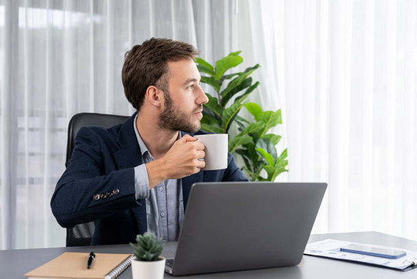 Businessman working on his laptop and drinking coffee at his desk in workspace. Smart executive taking coffee break after researching financial data and marketing plan on his corporate laptop. Entity - Foto, Imagen