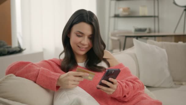 Hymyilevä nuori nainen, jolla on älypuhelin ja luottokortti, istuu sohvalla nykyaikaisessa kodissa. Happy naaras shopper käyttäen instant helppo mobiilimaksut, asiakas tekee ostoksia verkkokaupasta, e banking app - Materiaali, video