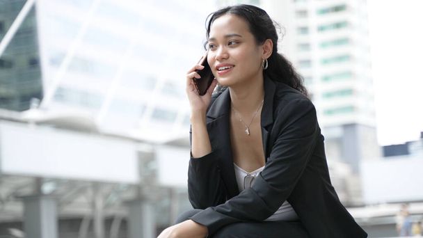 A young Asian businesswoman in formal attire sits smiling and using a smartphone, while listening to someone on the phone in Bangkoks bustling corporate office environment - Фото, зображення