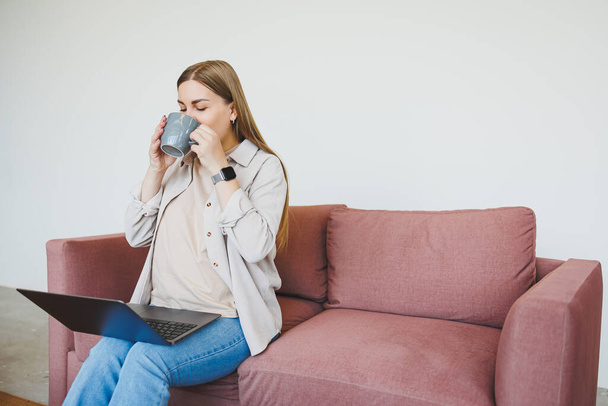 Smiling female freelancer in casual clothes sitting in comfortable sofa in modern cafeteria at table with laptop and phone, talking on mobile phone. Remote work - Photo, Image
