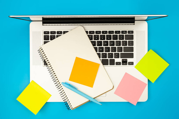 Laptop, notepad and colored paper reminders on blue background, flat lay, copy space. - Foto, imagen