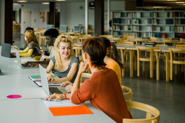 Group of multiracial students sitting at table with laptops books and conversing while female blonde smiling at unrecognizable male leaning on hand and looking at each other in library - Photo, Image