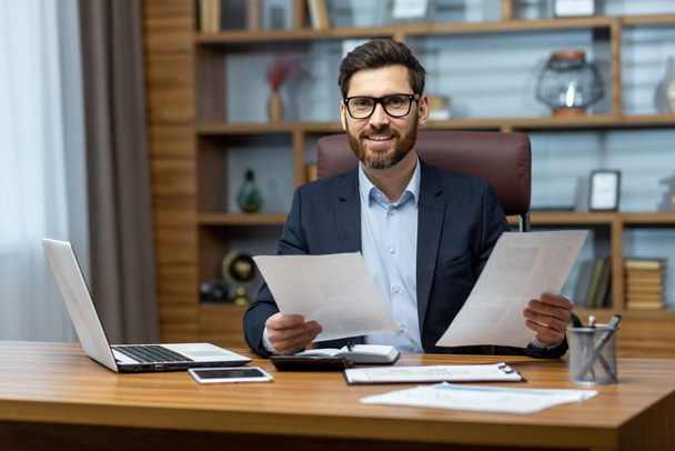 Portrait of successful financier, man with documents in hands smiling and looking at camera, businessman behind paper work sitting at desk inside office, mature boss in glasses and business suit. - Фото, изображение