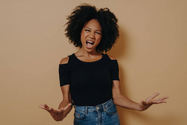 African woman in black t-shirt expressing anger, screaming with wide open mouth and palms open, isolated on beige studio background. Negative emotions concept. - Photo, Image