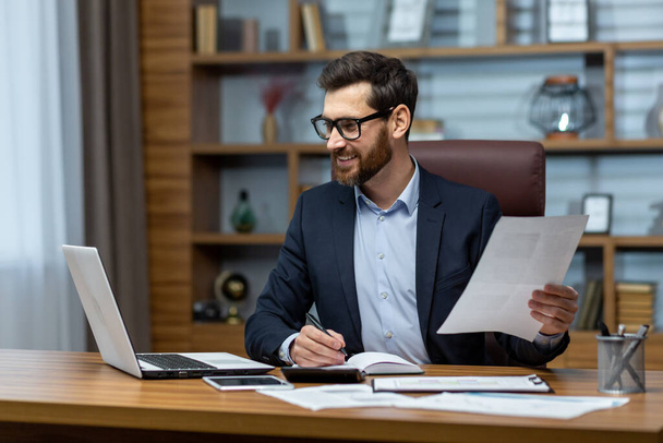 Mature businessman in shirt doing paperwork, man working with documents, contracts and bills sitting at table using laptop at work, financier accountant with beard and glasses - Foto, afbeelding