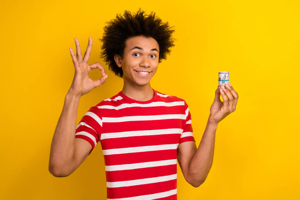 Foto van coole positieve man dragen gestreepte t-shirt tonen okey tonen geld cash roll geïsoleerde gele kleur achtergrond. - Foto, afbeelding