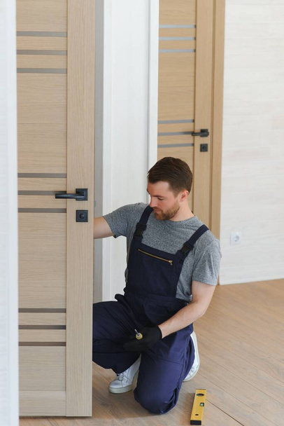 Installation of a lock on the front wooden entrance door. Portrait of young locksmith workman in blue uniform installing door knob. Professional repair service. Maintenance Concept. - Fotoğraf, Görsel