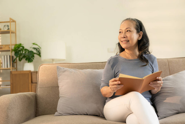 60 'lı yaşlarda Asyalı bir kadın, küçük oturma odasındaki kanepede kitap okuyan mutlu bir emekli kadın. Evinin rahatlığı. - Fotoğraf, Görsel