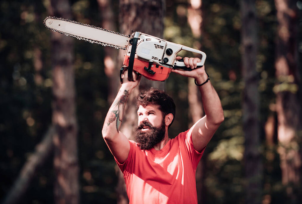 Stylish young man posing like lumberjack. Lumberjack on serious face carries chainsaw. Lumberjack worker standing in the forest with chainsaw. Man doing mans job - Fotografie, Obrázek
