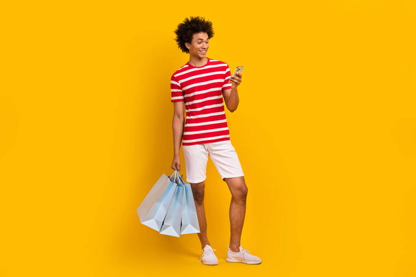 Full length photo of funny pretty man wear striped t-shirt holding shopper texting iphone gadget isolated yellow color background. - Photo, image