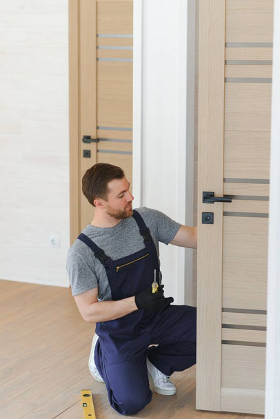 Installation of a lock on the front wooden entrance door. Portrait of young locksmith workman in blue uniform installing door knob. Professional repair service. Maintenance Concept. - Fotó, kép