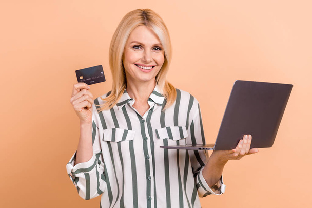 Portrait of positive satisfied retired woman striped formalwear hold laptop demonstrate credit card isolated on beige color background. - Fotografie, Obrázek