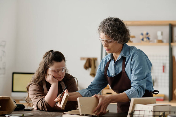 Carpenter in apron teaching girl to make birdhouse from wooden planks in workshop - Foto, imagen