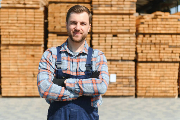 Portrait of a handsome worker choosing the best wooden boards. Carpenter standing next to a big stack of wood bars in a warehouse - Photo, image