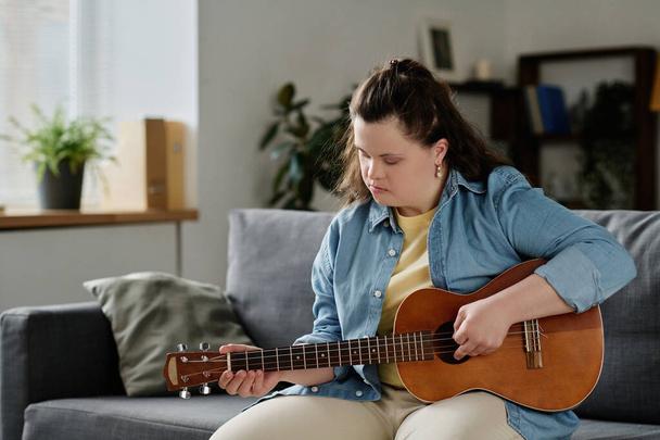 Chica con síndrome de Down aprender a tocar la guitarra mientras está sentado en el sofá en la sala de estar - Foto, imagen