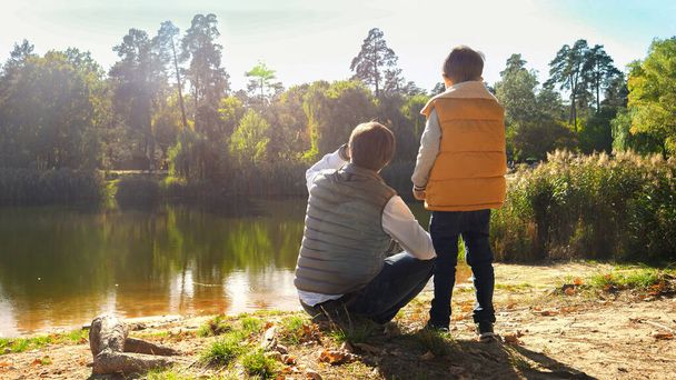 Маленький мальчик с маленьким отцом стоит у озера и смотрит на воду. Семья на открытом воздухе, счастливое воспитание и детство, осенний пейзаж, дети и родители отдыхают на улице - Фото, изображение