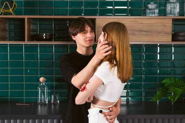Νεαρό ζευγάρι εφήβων στην κουζίνα, ο τύπος αγκαλιάζει το κορίτσι και προσπαθεί να ζητήσει συγγνώμη, αλλά το κορίτσι προσβάλλεται - Φωτογραφία, εικόνα