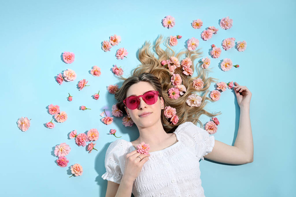 Beutiful blond woman lying with many pink roses on light blue background - Foto, afbeelding
